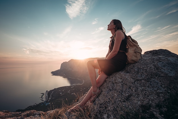 Una ragazza si siede sul bordo della scogliera e guarda il sole mare e montagne. L'idea e il concetto di ricreazione, libertà, estate, attività all'aperto e viaggi