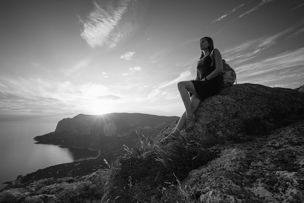 Una ragazza si siede sul bordo della scogliera e guarda il sole mare e montagne. L'idea e il concetto di ricreazione, libertà, estate, attività all'aperto e viaggi