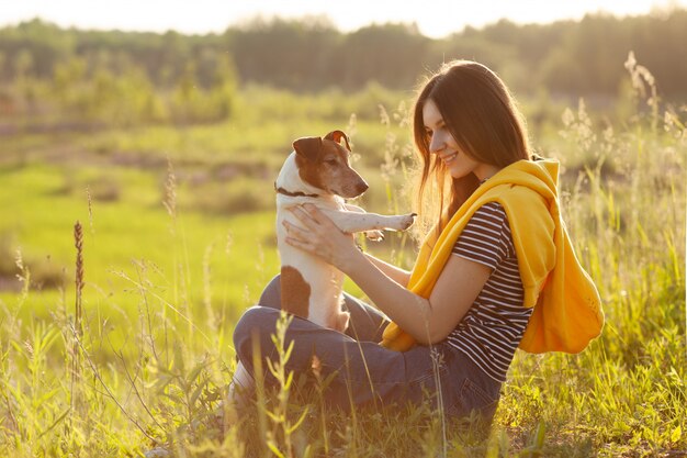 Una ragazza si siede sull'erba sotto i raggi del sole al tramonto e tiene il suo cane in braccio.