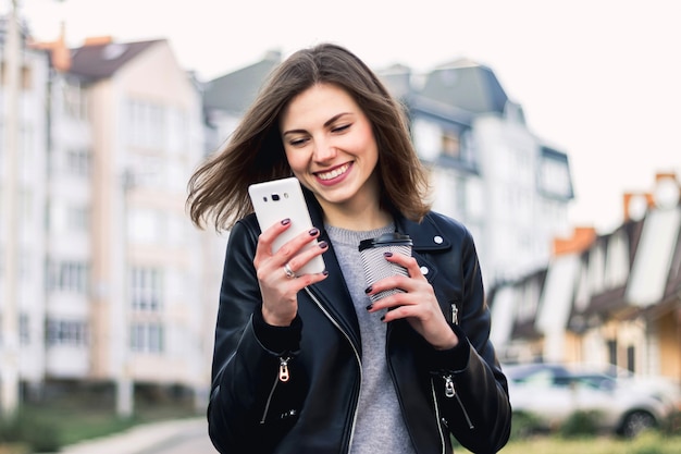 Una ragazza sorride e cammina per la città con un telefono cellulare e una tazza di caffè