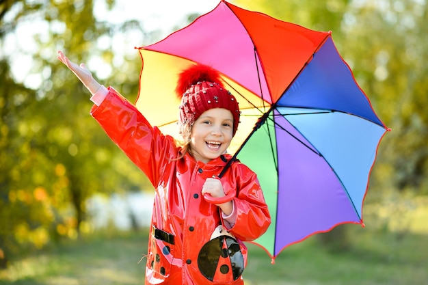 Una ragazza sorridente con un cappello rosso e una giacca rossa cammina con un ombrello arcobaleno in natura