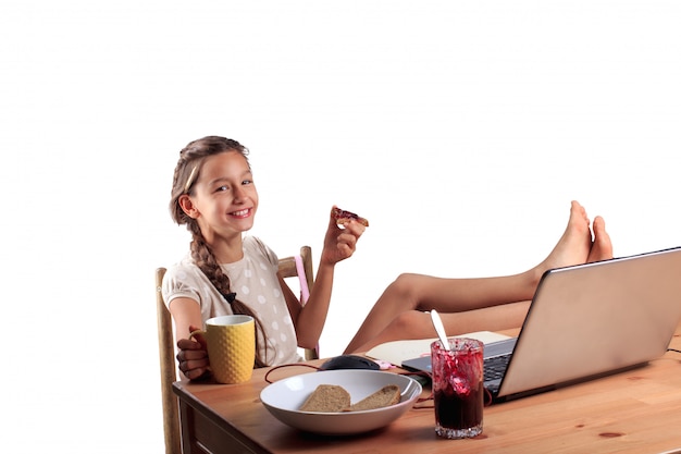 Una ragazza sorridente felice con un espressivo volto emotivo seduto al tavolo con un computer portatile, mangiando pane con marmellata e tenendo una tazza di tè isolata