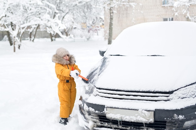 Una ragazza spazza la neve dal cofano di un'auto con una spazzola dopo una forte nevicata