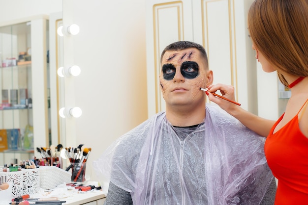 Una ragazza specialista del trucco fa un trucco festivo e raccapricciante per un uomo per le vacanze di Halloween in studio. Felice concetto di Halloween.