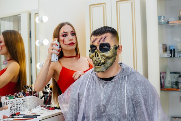 Una ragazza specialista del trucco fa un trucco festivo e raccapricciante per un uomo per le vacanze di Halloween in studio. Felice concetto di Halloween.