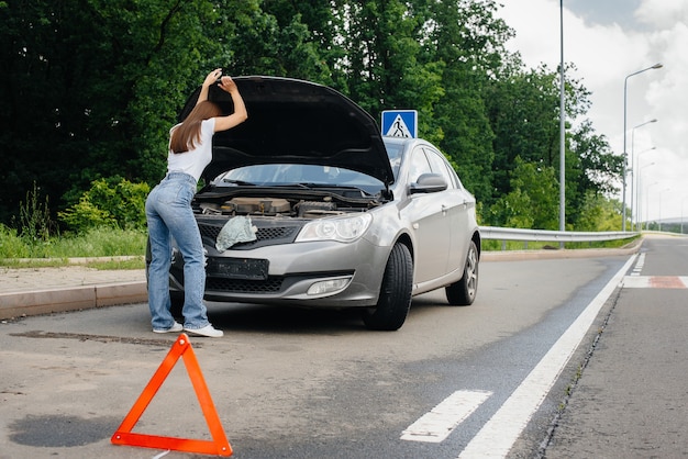 Una ragazza sta vicino a un'auto in panne nel mezzo dell'autostrada e guarda sotto il cofano. Guasto e guasto dell'auto. In attesa di aiuto.