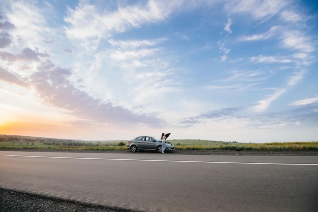 Una ragazza sta vicino a un'auto rotta nel mezzo dell'autostrada durante il tramonto e cerca di ripararla. Riparazione e riparazione dell'auto. Risoluzione dei problemi.