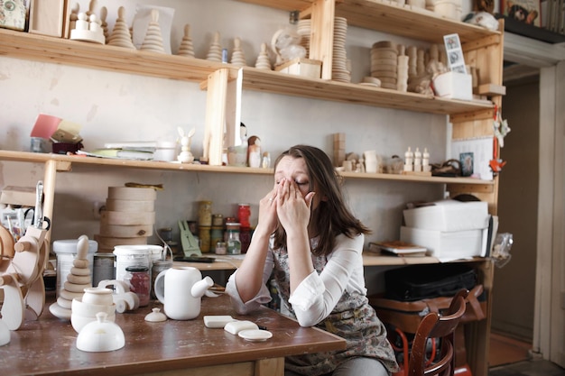 Una ragazza stanca si strofina gli occhi con le mani mentre è seduta a un tavolo in un laboratorio d'arte che produce giocattoli di legno