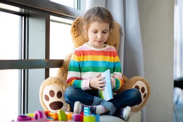 Una ragazza sul divano vicino alla finestra con un grande orso giocattolo e legge un libro Kids Teaching