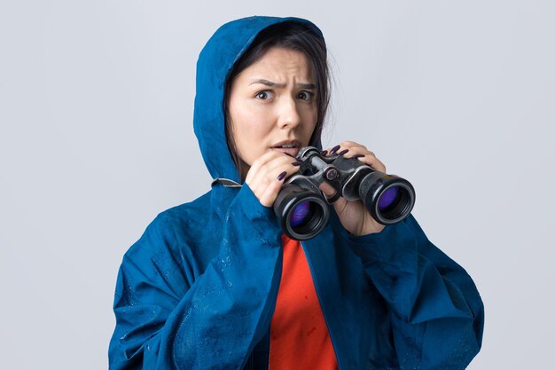 Una ragazza turistica con un impermeabile blu tiene in mano un binocolo e guarda lontano, spie.