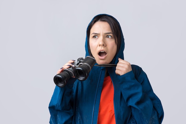 Una ragazza turistica con un impermeabile blu tiene in mano un binocolo e guarda lontano, spie.