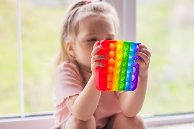 Una ragazzina bionda si siede vicino alla finestra e gioca con il nuovo giocattolo sensoriale di tendenza: arcobaleno pop it. Antistress semplice fossetta giocattolo olorful. Squishy giocattoli a bolle morbide color arcobaleno