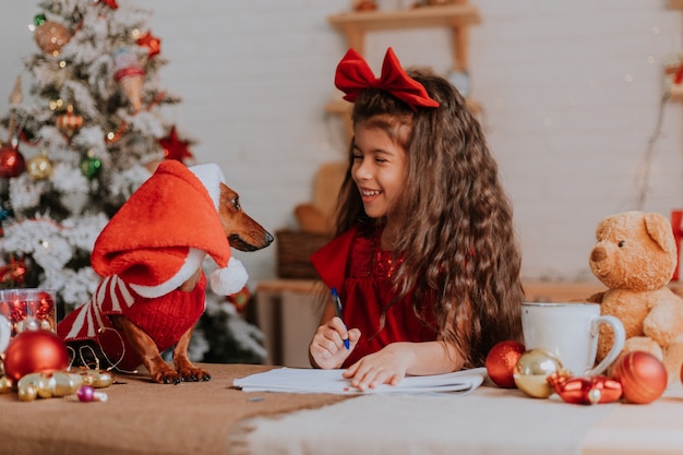 Una ragazzina carina con un bassotto nano scrive una lettera con gli auguri al concetto di capodanno di Babbo Natale