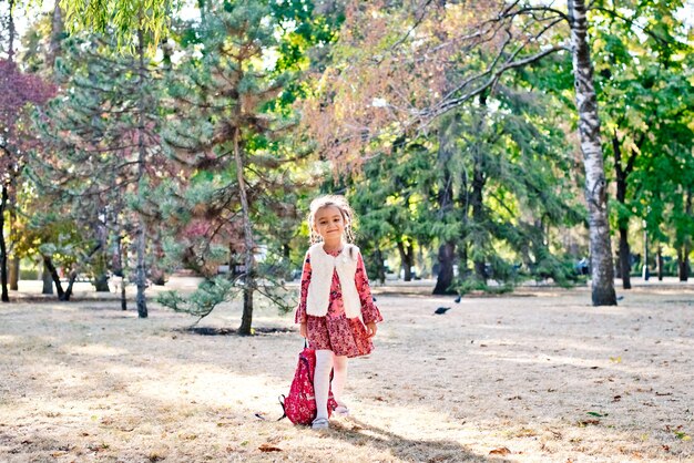 una ragazzina carina con un vestito rosso una studentessa cammina in un parco autunnale con uno zaino