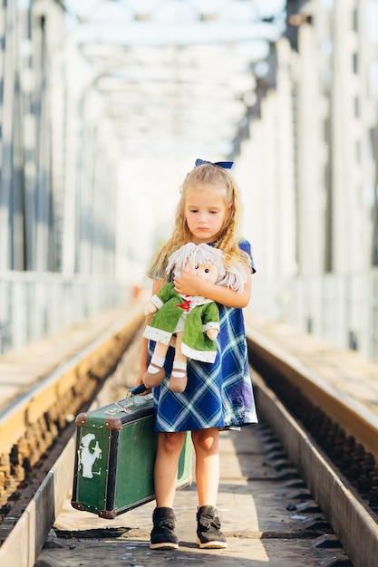 Una ragazzina carina con un vestito sta aspettando il suo treno con una valigia alle stazioni ferroviarie