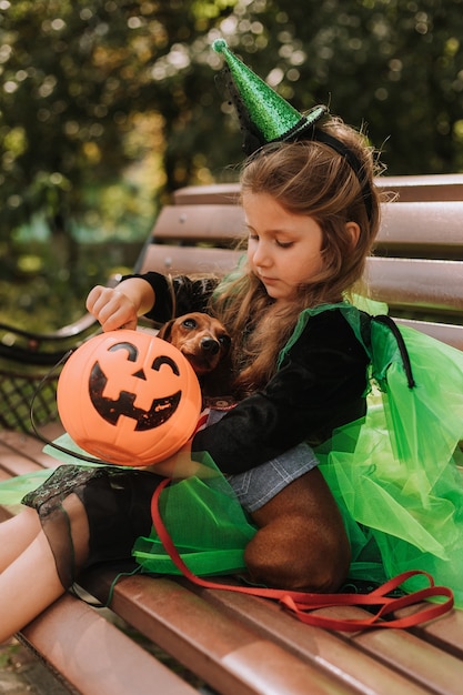 Una ragazzina carina con una strega verde di Halloween o un costume da fata è seduta su una panchina per strada