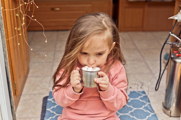 Una ragazzina carina si siede sui gradini di una roulotte e beve cioccolata calda con marshmallow