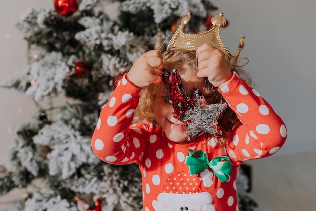 Una ragazzina dai capelli ricci in un vestito di carnevale nascondeva il viso dietro le lucide stelle dei giocattoli dell'albero di Natale