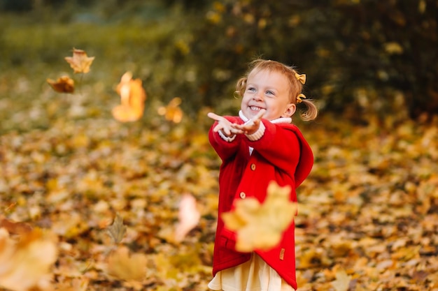 Una ragazzina divertente con un cappotto rosso cattura le foglie autunnali divertendosi e camminando nella foresta autunnale