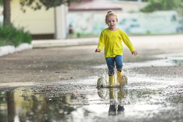 una ragazzina in abiti gialli e stivali di gomma corre allegramente attraverso le pozzanghere dopo la pioggia