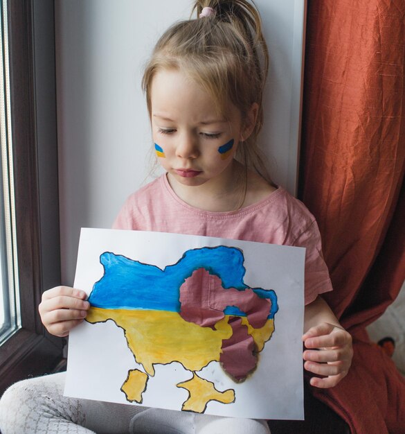 Una ragazzina ucraina con una faccia triste è seduta sul davanzale della finestra con in mano una mappa bruciata dell'Ucraina con grandi buchi nelle sue mani Bambini e guerra