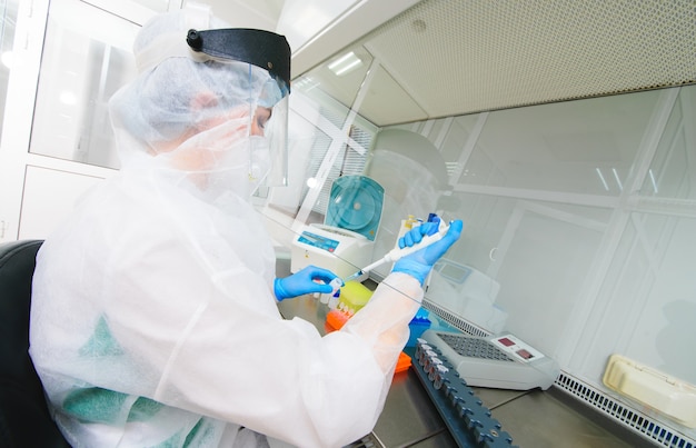 Una ricercatrice di laboratorio PCR femminile in tuta medica protettiva, maschera sterile, casco, guanti di gomma con un erogatore in mano conduce un esperimento scientifico o sviluppa un vaccino.