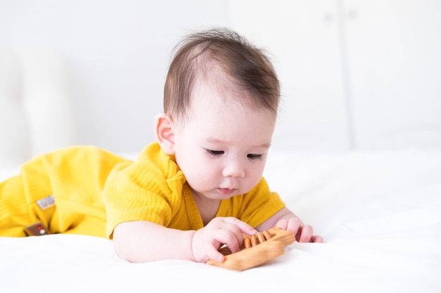 una sana bambina asiatica in tuta gialla gioca con un massaggiagengive giocattolo in legno per denti su biancheria da letto bianca