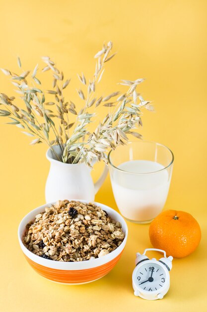 Una sana colazione abbondante. Muesli al forno in una ciotola, un bicchiere di latte, arancia e sveglia sulla superficie gialla. Vista verticale