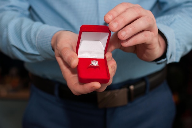 Una scatola aperta di velluto rosso con un anello in oro bianco con una pietra preziosa nelle mani di un uomo con una camicia blu