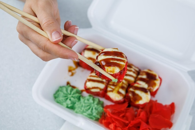 Una serie di rotoli giapponesi in una scatola di plastica su un tavolo luminoso La ragazza prende il cibo con le bacchette Sushi a consegna rapida in un contenitore bianco