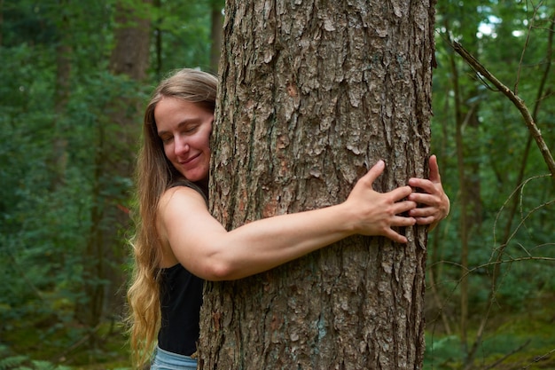 Una signora bionda caucasica con i capelli lunghi che abbraccia un albero nella foresta