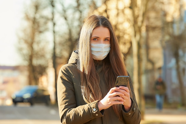 Una signora che utilizza uno smartphone indossa una maschera medica per evitare la diffusione del coronavirus in una strada cittadina. Una ragazza con una maschera chirurgica sul viso contro COVID-19.