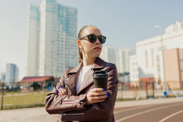 Una signora elegante e indipendente con i capelli scuri che indossa occhiali da sole e una giacca bordeaux sta guardando da parte mentre cammina in città con un caffè alla luce del sole