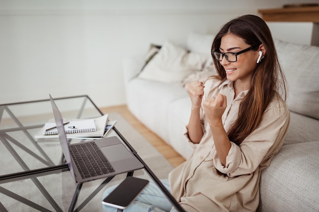 Una signora europea millenaria soddisfatta con gli occhiali guarda il computer e fa un gesto di vittoria con le mani