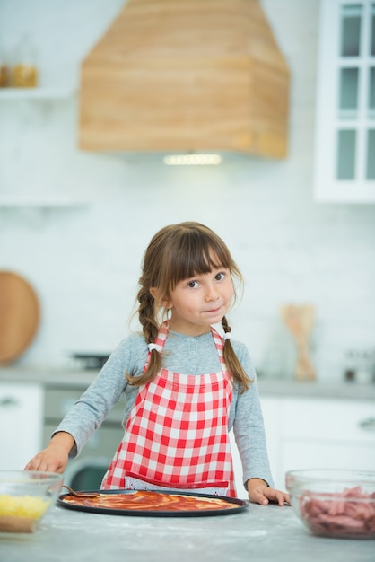 Una simpatica bambina con le trecce in un grembiule a scacchi prepara la pizza da sola in cucina. Corso di cucina per bambini.