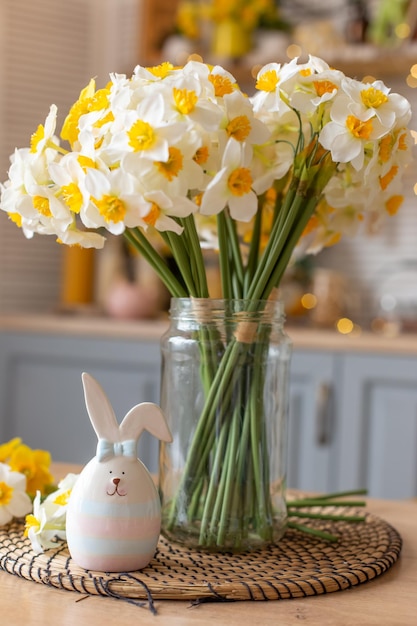 Una simpatica statuetta in ceramica di coniglietto si trova sul tavolo della cucina accanto a un vaso di vetro con un flusso di narcisi