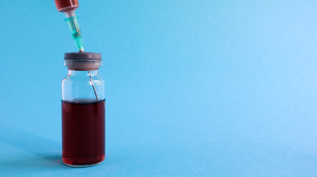 Una siringa sporge da una bottiglia di liquido rosso. Isolato su sfondo blu. Medicina, iniezioni, vaccini e siringhe usa e getta, concetto di droga. Bottiglia sterile. Fiala di vetro medica per iniezione.