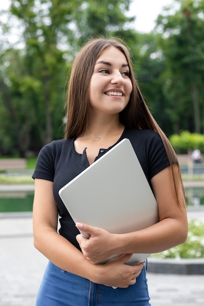 Una sorridente ragazza europea dai capelli scuri in abiti casual si trova in un parco in estate abbracciando il suo laptop