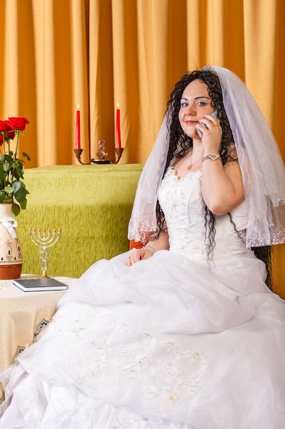 Una sposa ebrea in abito da sposa bianco con velo si siede a un tavolo con fiori e parla al telefono prima della cerimonia chuppah. Foto verticale