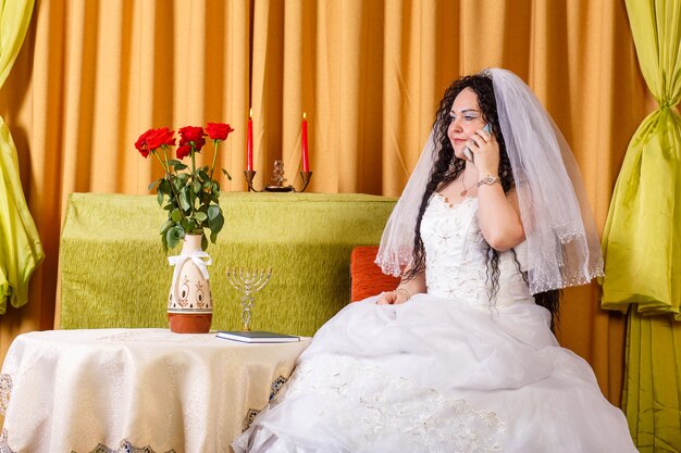 Una sposa ebrea in abito da sposa bianco con velo si siede a un tavolo con fiori e parla al telefono prima della cerimonia chuppah