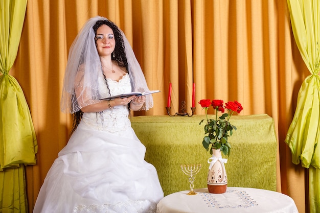 Una sposa ebrea in abito da sposa bianco con velo si trova nella hall a un tavolo con fiori e prega prima della cerimonia chuppah