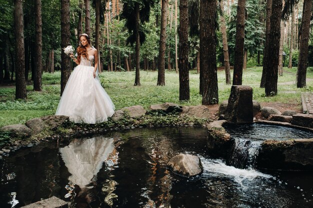 Una sposa elegante in un abito bianco e guanti che tengono un bouquet si erge vicino a un ruscello nella foresta, godendosi la natura. Un modello in abito da sposa e guanti in un parco naturale. Bielorussia