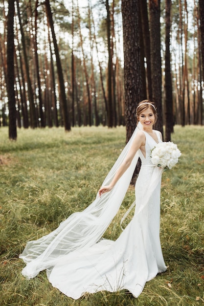 Una sposa elegante si trova nella foresta e tiene in mano un bellissimo bouquet da sposa con una peonia