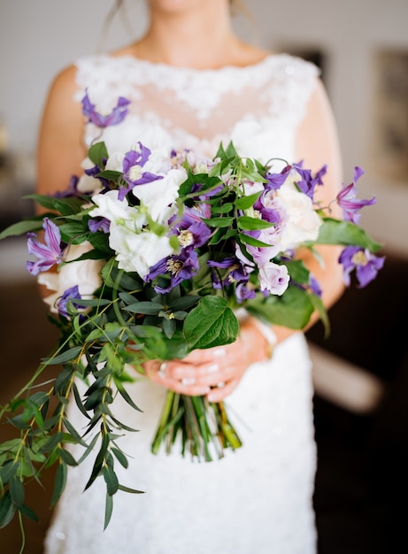Una sposa in un abito di pizzo tiene un insolito bouquet con rose clematidi viola e rami verdi