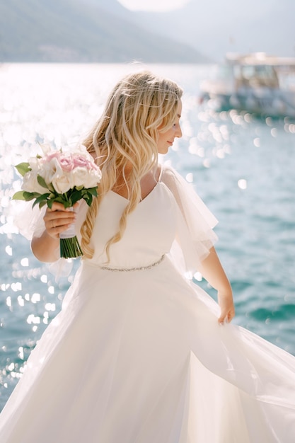 Una sposa in un vestito delicato con un bouquet in mano si trova su un molo nella baia di Kotor