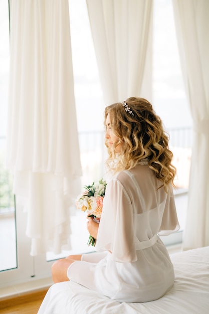 Una sposa in una vestaglia delicata con un bouquet in mano seduta sul letto vicino alla finestra con il bianco