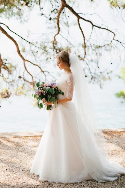 una sposa sofisticata sta con un bouquet da sposa tra le mani sotto i rami di un albero