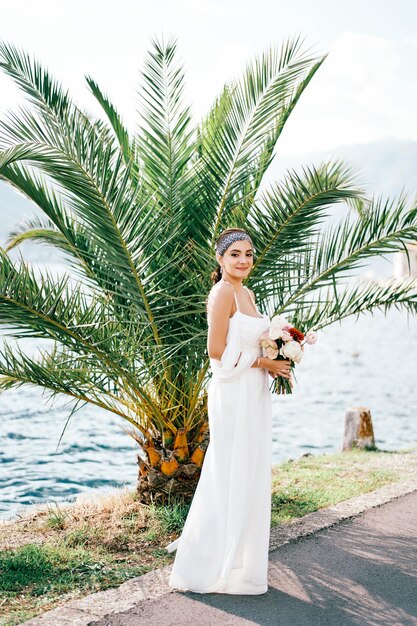 Una sposa sorridente con un bouquet in piedi vicino a una palma sulla riva del mare