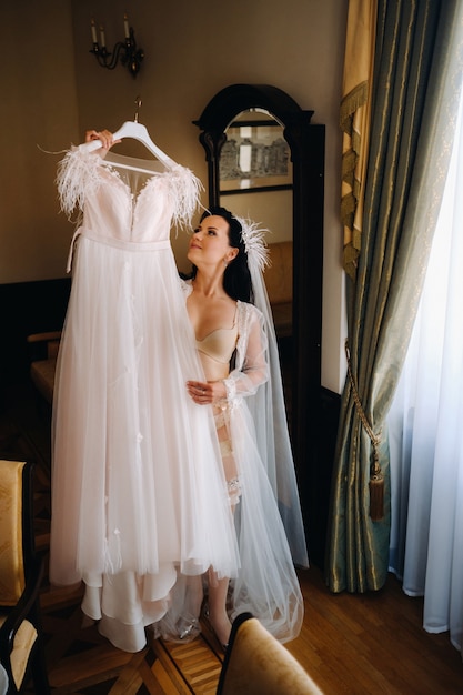 Una sposa vestita con un abito trasparente da boudoir e biancheria intima tiene il suo abito da sposa tra le mani all'interno della casa.