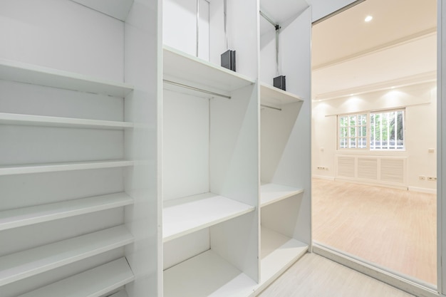 Una stanza vuota con pareti dipinte di bianco con accesso a uno spogliatoio con più scaffali marera bianchi e finestre in alluminio in stile francese